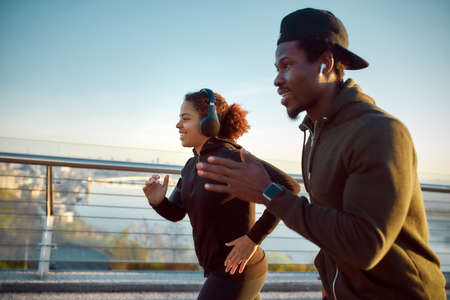 Morning run. Side view of happy sporty African couple in headphones jogging together on the bridge...