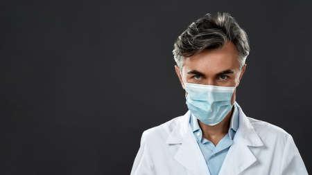 Protective equipment. Confident mature male doctor wearing medical uniform and protective mask looking at camera while standing against black backgroundの写真素材