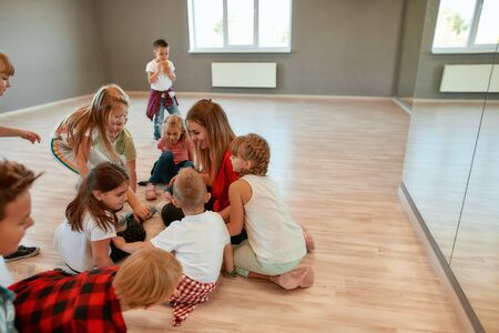 Spending time with kids. Full length portrait of positive dance teacher talking to group of little girls and boys sitting on the floor in studioの写真素材