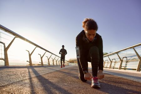 Getting ready for jogging. African sporty woman in black sportswear tying shoelaces before run with...