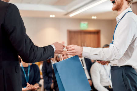 Your reward. Cropped shot of young male speaker receiving gift bag from his male colleague at business meeting, forumの写真素材