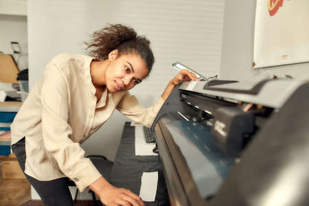 Checking the process. Young woman controlling, monitoring printing machine while printing advertisement sticker with text or picture for a T-shirtの写真素材