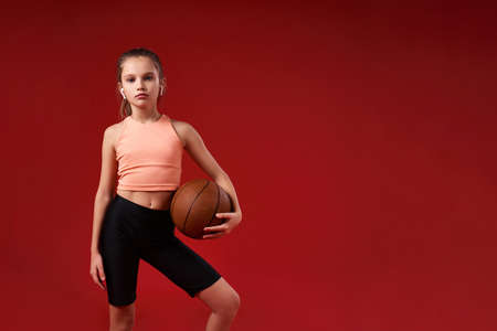 Ready to play. A cute kid, girl is engaged in sport, looking at camera while exercising with basketball. Isolated on red background. Fitness, training, active lifestyle concept. Horizontal shotの写真素材