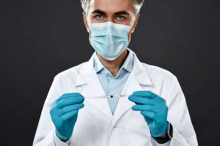 Mature doctor or scientist in uniform and medical mask holding syringe with vaccine from flu, coronavirus, covid-19 while standing against black backgroundの写真素材