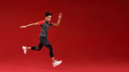 Just run. A teenage boy is engaged in sport, he is looking down while jumping. Isolated on red background. Fitness, training, active lifestyle concept. Horizontal shotの写真素材