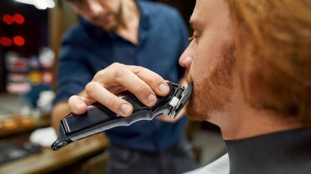 Handsome redhead guy sitting in armchair in the barbershop while barber trimming his mustache. Working with electric clipper machineの写真素材