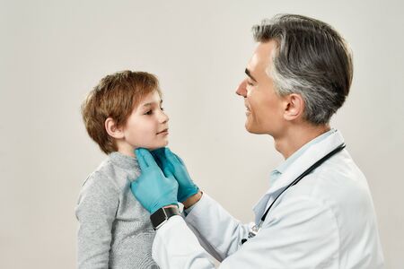 Sore throat. Professional mature pediatrician or doctor examining thyroid glands of a little boy while standing against grey backgroundの写真素材