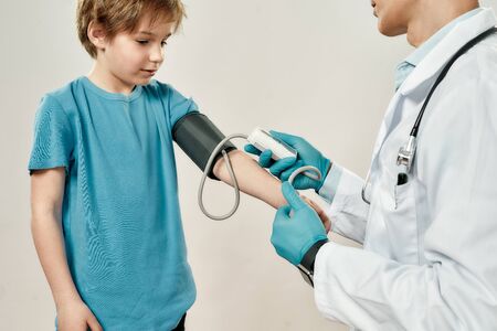 Pediatric specialist working with child. Professional doctor in medical uniform and sterile gloves measuring blood pressure of cute little boy, standing against grey backgroundの写真素材