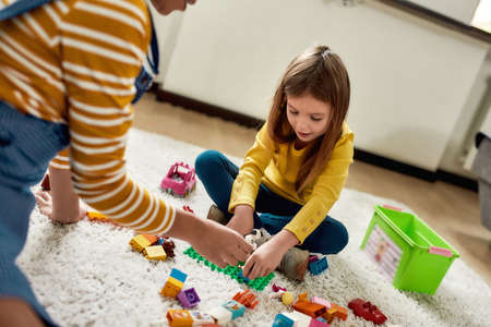 Game. Caucasian cute little girl spending time with african american baby sitter. They are playing with toys, sitting on the floorの写真素材
