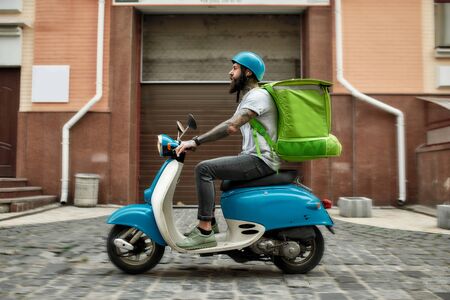 Faster is always better. Bearded delivery man in helmet with thermo bag or backpack riding a motor scooter along the city, delivering food. Courier, delivery service conceptの写真素材