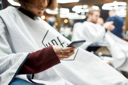 Chatting with a friend. Cropped photo of a young guy using mobile phone during a haircut in the...