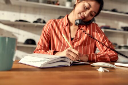 Portrait of young female worker receiving orders online or via telephone while sitting in the office. Woman working at custom T-shirt, clothing printing companyの写真素材