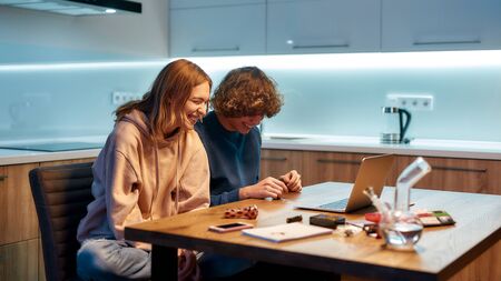Happiness drug. Happy couple looking at laptop screen and laughing while sitting in the kitchen at...