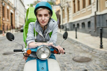 Young woman in helmet with thermo backpack looking at camera, delivering food on a motor scooterの写真素材