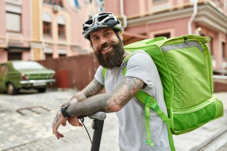 Ready, set, delivered. Brutal bearded delivery man in helmet with thermo backpack smiling at camera while riding an electric scooter. Courier, delivery service conceptの写真素材