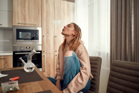 Relieve stress. Young caucasian woman exhaling the smoke while smoking marijuana from a bong or glass water pipe, sitting in the kitchen. Buds in a plastic bag and lighter on the tableの写真素材