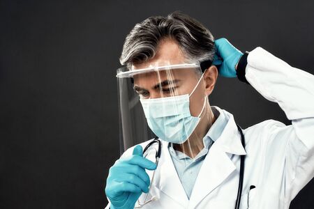 Close up portrait of mature male doctor in medical mask and blue gloves wearing face shield or protective gear while standing against dark backgroundの写真素材