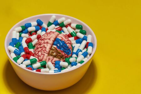 Creative concept with different medical pills, capsules lying in bowl with model of human brain on yellow background. Health care and treatment conceptの写真素材