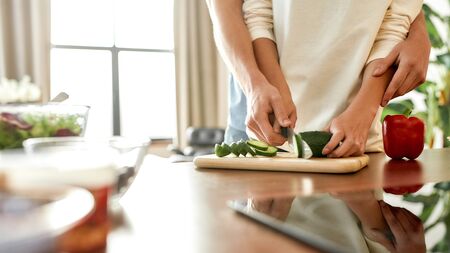 Cropped shot of woman cutting vegetables while man helping, holding her hands, standing behind her. Vegetarians preparing healthy meal in the kitchen together. Focus on handsの写真素材
