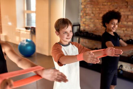 Caucasian boy working out using resistance band together with other kids in gym. Sport, healthy lifestyle, physical education conceptの写真素材