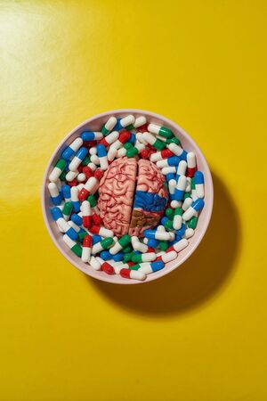 Creative concept with different medical pills, capsules lying in bowl with model of human brain on yellow background. Health care and treatment conceptの写真素材