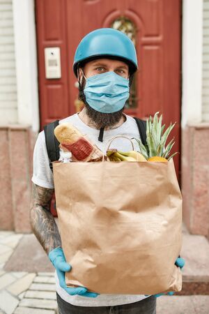 Bearded delivery man in helmet wearing mask and gloves due to the emergence of the Covid19 virus, holding grocery bag, while delivering food. Courier, delivery service, lockdown conceptの写真素材