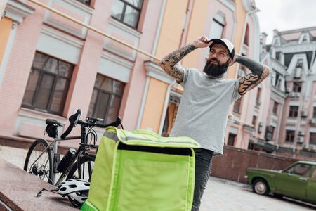 Always on time. Brutal stylish delivery man in cap looking aside, standing with backpack outdoors,...
