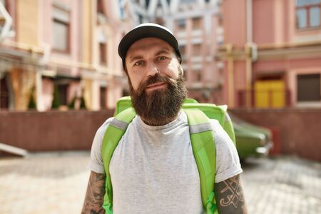 Delivery matters. Brutal bearded delivery man in cap looking at camera, standing with backpack outdoors, ready to work. Courier, delivery service conceptの写真素材