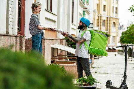 Bearded delivery man in helmet with thermo backpack giving away pizza to a female customer, while delivering food. Courier, delivery service conceptの写真素材