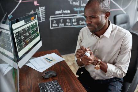 Money is required. Stylish african businessman, trader sitting by desk in front of multiple...