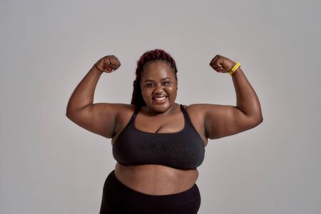 Enjoy your body. Plump, plus size african american woman in sportswear smiling at camera, showing strong arms in studio over grey background. Concept of sport, healthy lifestyle, body positiveの写真素材