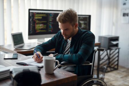 Side view of young concentrated male web developer in a wheelchair making some notes while sitting...