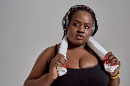 Results with resolve. Plump, plus size african american woman in sportswear and headphones holding towel around her neck, posing in studio over grey backgroundの写真素材
