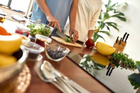 Cropped shot of man cutting vegetables while woman holding pepper and standing near him. Vegetarians preparing healthy meal in the kitchen together. Focus on cucumber and cutting board. Dutch angleの写真素材