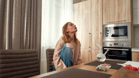 Take it easy. Young caucasian woman exhaling the smoke while smoking marijuana from a bong or glass water pipe, sitting in the kitchen. Red marijuana grinder and cannabis buds on the tableの写真素材