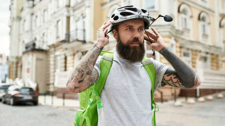 Helping you. Brutal bearded delivery man wearing helmet while standing with thermo bag or backpack...