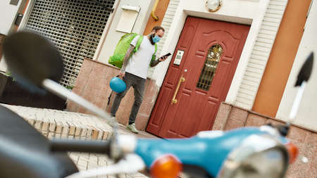 Full length shot of bearded delivery man in mask and gloves with thermo backpack using smartphone, standing by the door, while completing the order. Courier, delivery service conceptの写真素材