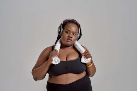 Plump, plus size african american woman in sportswear and headphones holding towel around her neck,...