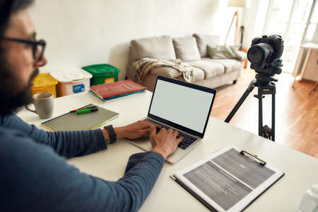 Side view of male blogger wearing eyeglasses using laptop while recording video blog on professional digital equipment at homeの写真素材