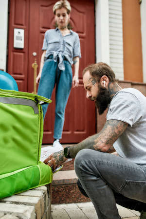 Bearded delivery man taking pizza out of thermo backpack while giving away order to a female customer. Courier, delivery service conceptの写真素材