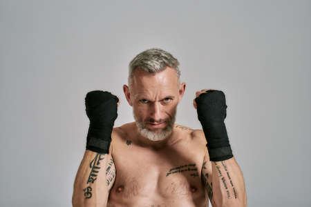 Refuse to Lose. Half naked middle aged athletic man, kickboxer looking angry holding hands standing in studio over grey background. Muay Thai, Boxing or Kickboxing conceptの写真素材