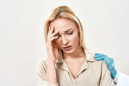 Portrait of depressed young woman visiting doctor. Doctor in blue sterile gloves supporting and...