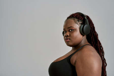 Plump, plus size african american woman in sportswear and headphones looking at camera, standing in studio over grey background. Concept of sport, healthy lifestyle, body positive, equalityの写真素材