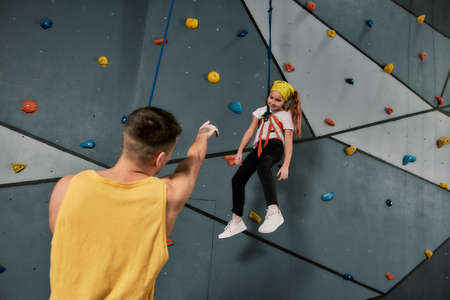 Male instructor holding rope, talking to a girl in safety equipment and harness while she is...