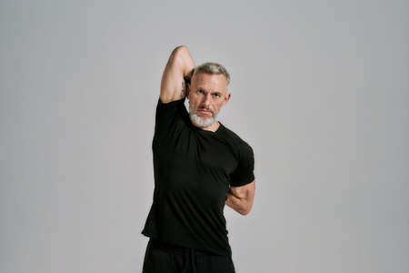 Middle aged muscular man in black sportswear looking at camera while warming up his body before...