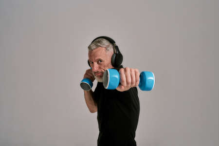 Middle aged muscular man in black t shirt and headphones looking at camera holding blue dumbbells,...