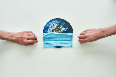 Conceptual flatlay with hands holding medical mask on planet Earth made of paper over white...