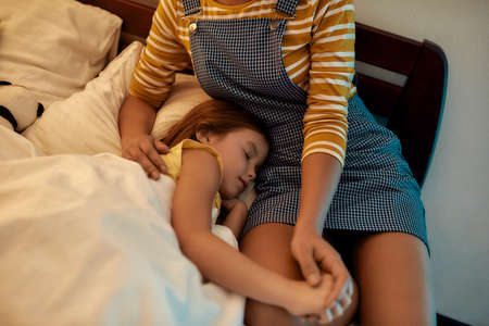 Your kid in lovable hands. African american woman baby sitter spending time with little girl in the evening. Nanny sitting with kid, watching her falling asleep in bedの写真素材