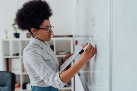 Online english course. Side view of a young afro american female teacher writing on a whiteboard while teaching English language via internet from homeの写真素材