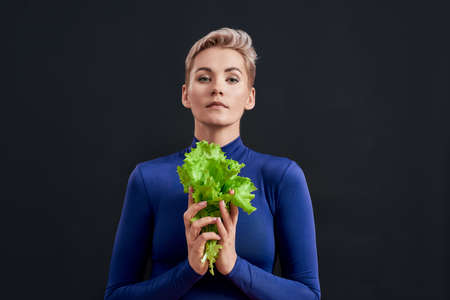 Portrait of attractive woman with pierced nose and short hair in blue turtleneck looking at camera, holding fresh green lettuce isolated over dark backgroundの写真素材